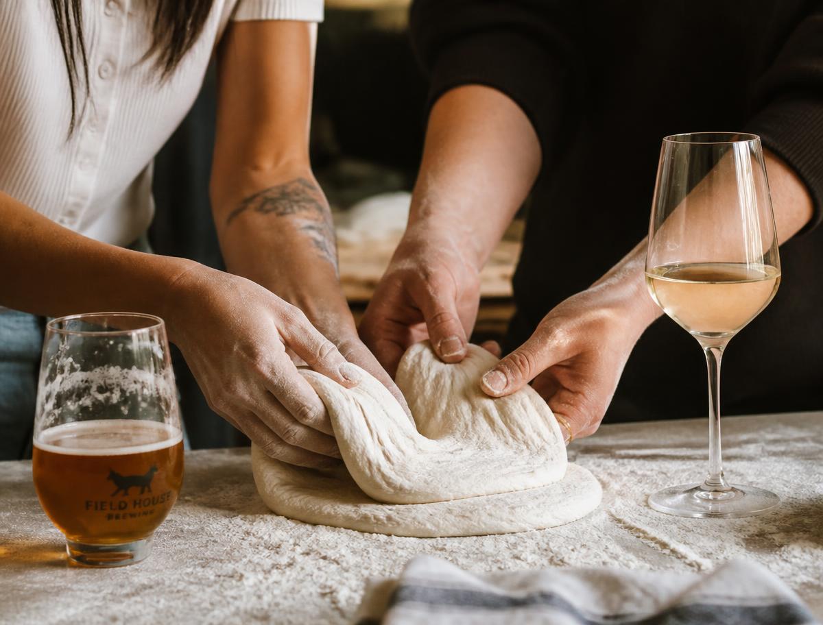 Two people stretching and folding sourdough dough together beside a glass of wine and a craft beer